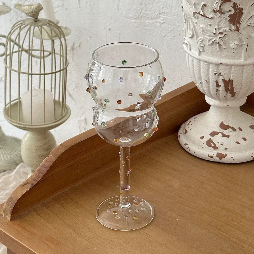 Decorative glass with embellishments on a wooden surface with a birdcage and candle holder in the background.
