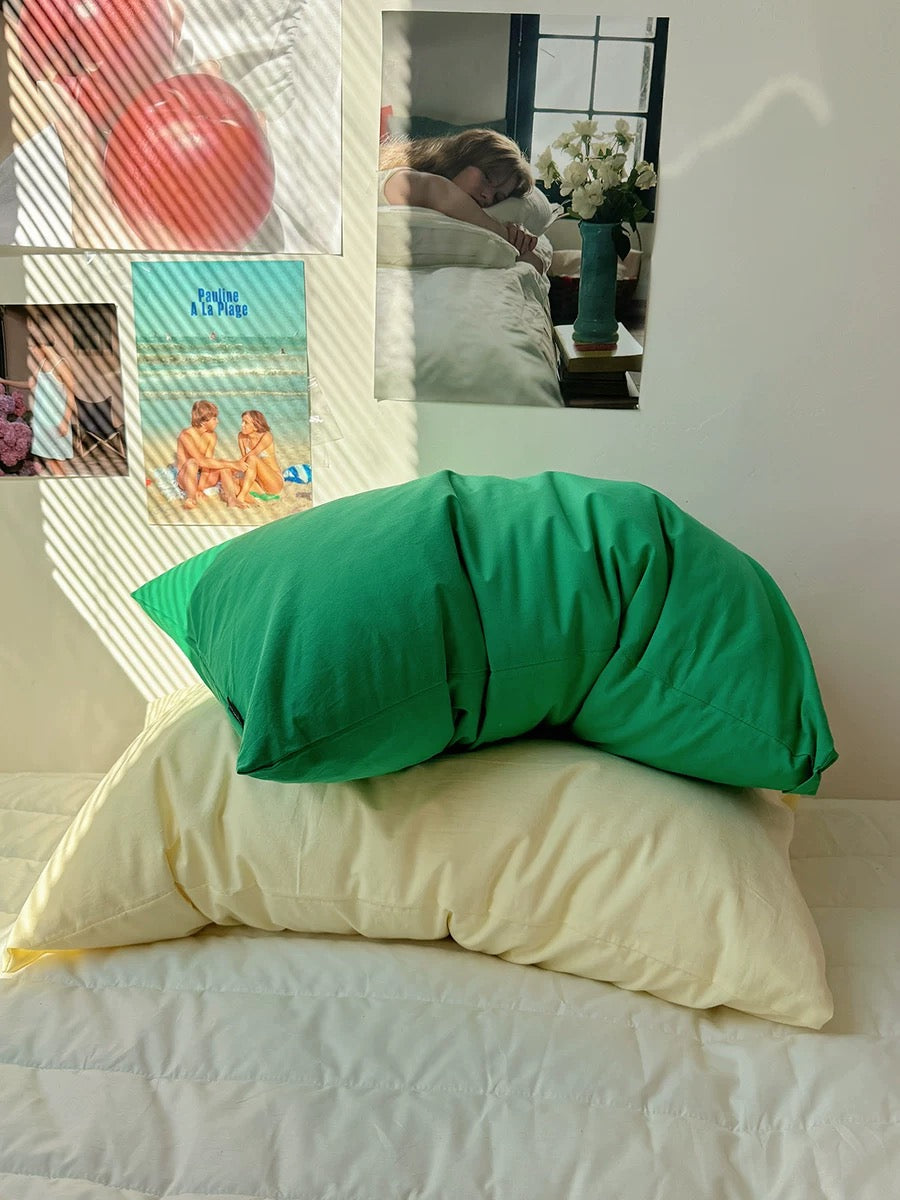A bright green pillowcase-shaped pillow resting on top of a cream-colored pillow on a bed, with a wall collage in the background.
