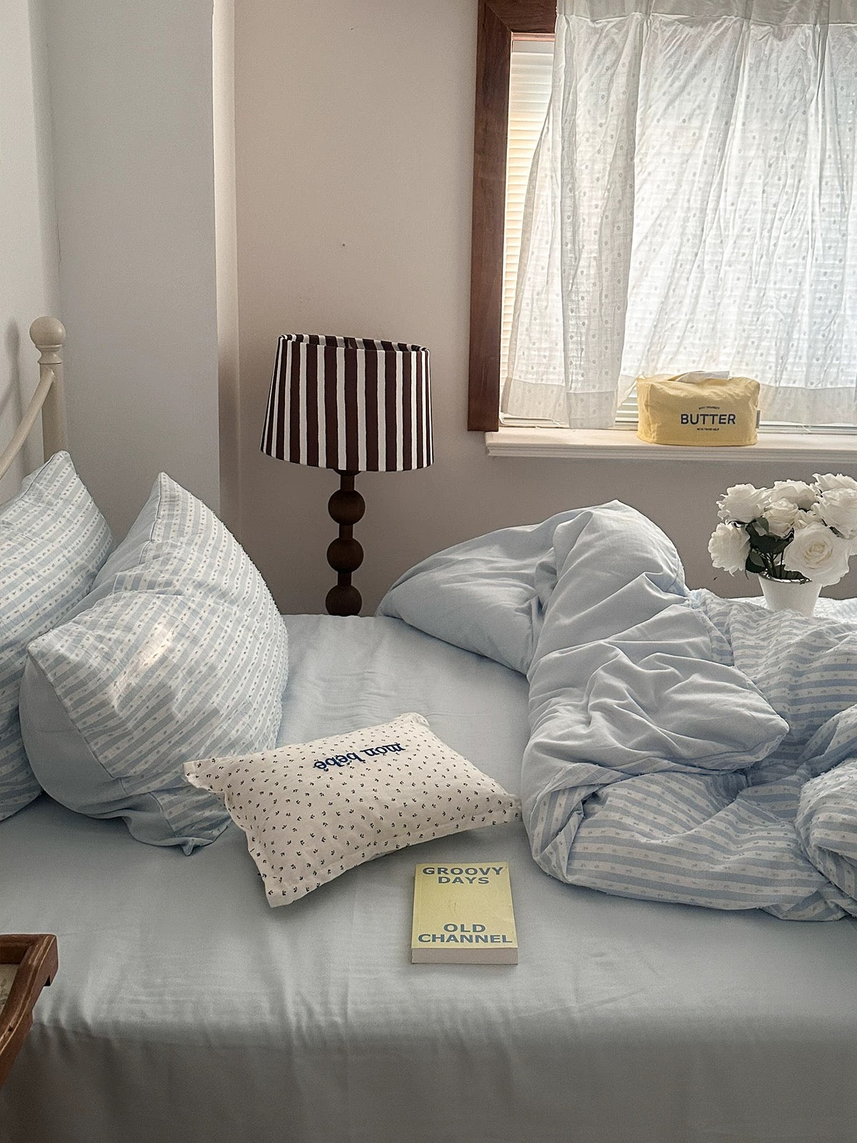 A styled bedroom scene showing a bed made with light blue and white striped bedding, including a duvet cover, pillowcases, and a small decorative pillow. A striped table lamp, a window with sheer curtains, and a vase of white flowers are visible in the room.