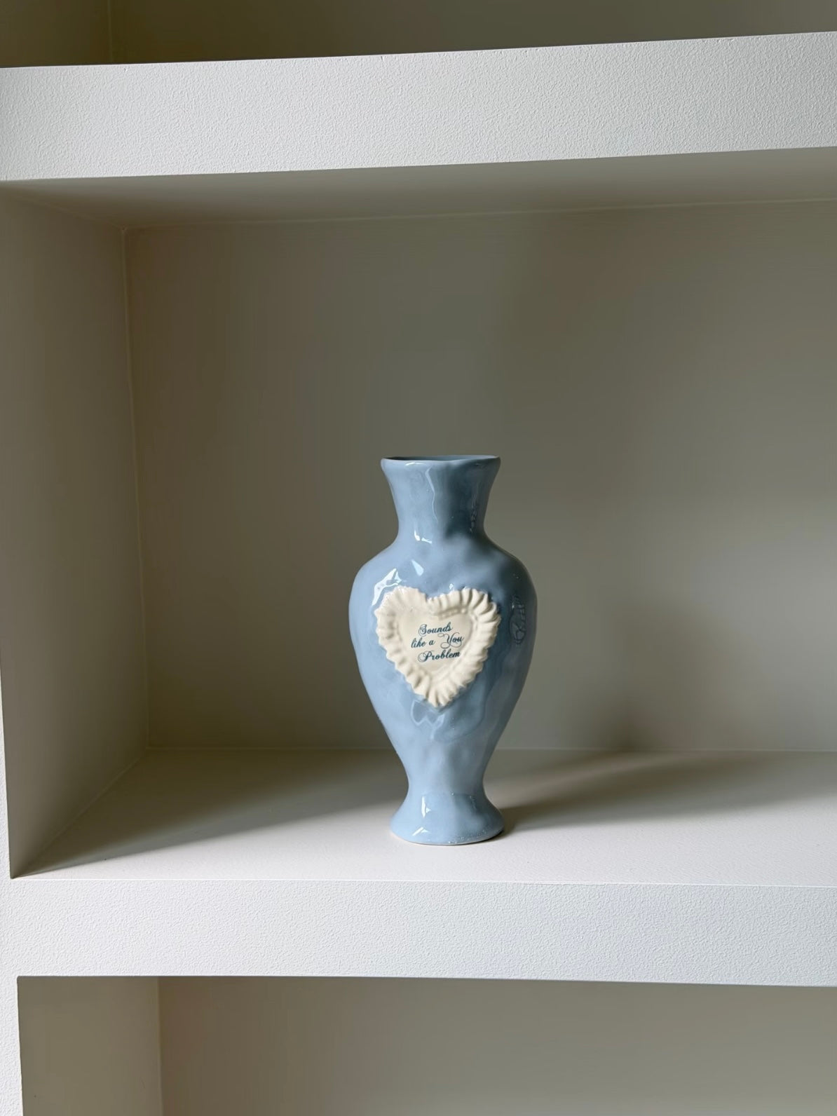 Coquette blue vase on a white shelf, with curved shoulders, a narrow neck and a raised ruffled heart on the front.
