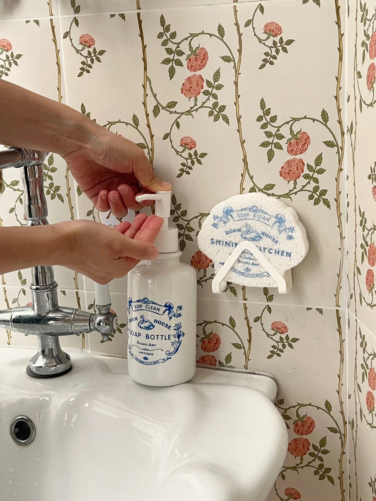 Coquette soap dispenser with blue swan artwork stands beside a faucet as hands press the white pump.