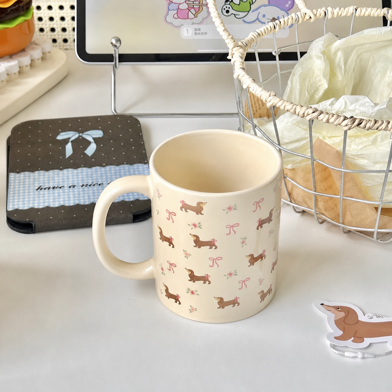 A cream-colored ceramic mug with a pattern of brown dachshunds and pink bows, set on a white desk next to a tablet, a phone case, and a wire basket.
