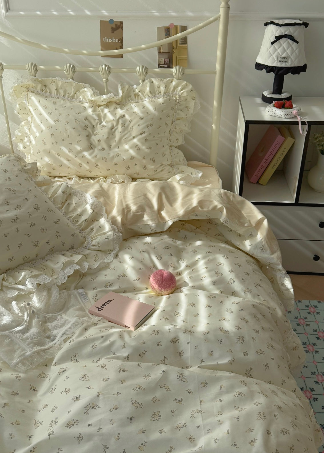 Bed with white floral bedding, pink notebook, and pink flower on a white nightstand with books and lamp.