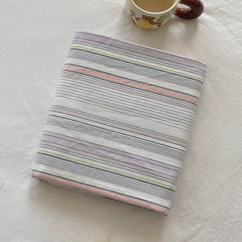 A folded striped fabric item, likely a blanket or quilt, with pastel horizontal stripes in lavender, pink, blue, and yellow on a white background. A ceramic mug is partially visible at the top edge.