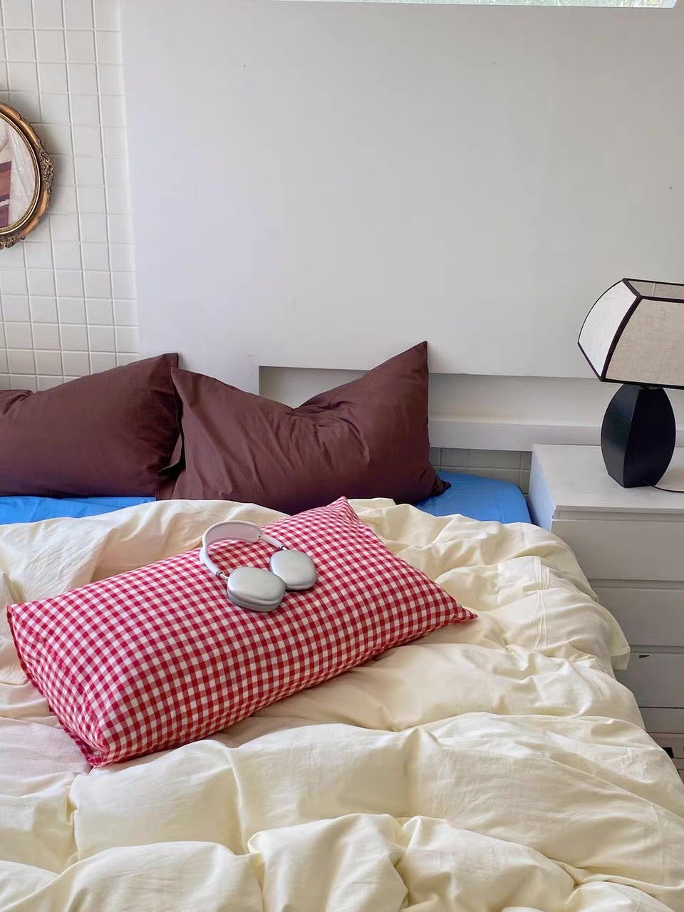 A styled bedroom scene showing a bed with cream bedding, two dark brown pillows, a red-and-white gingham accent pillow, and a pair of white slippers on top. A white bedside table with a black lamp is visible on the right.