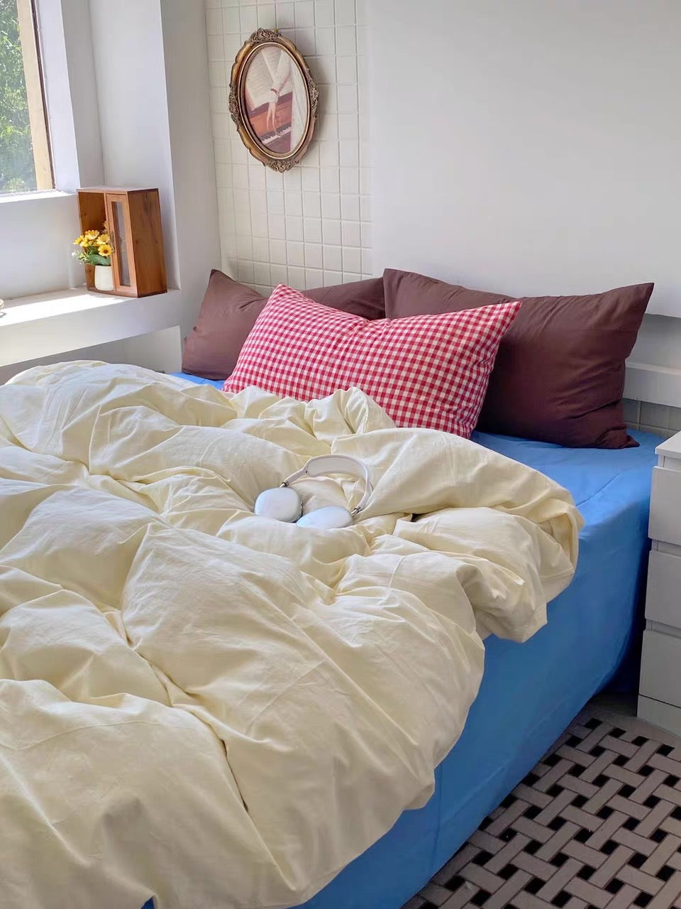 A cozy bedroom scene with a bed made up in soft cream bedding, a bright blue sheet, and several pillows including a red-and-white gingham accent pillow. A pair of white over-ear headphones rests on the duvet.