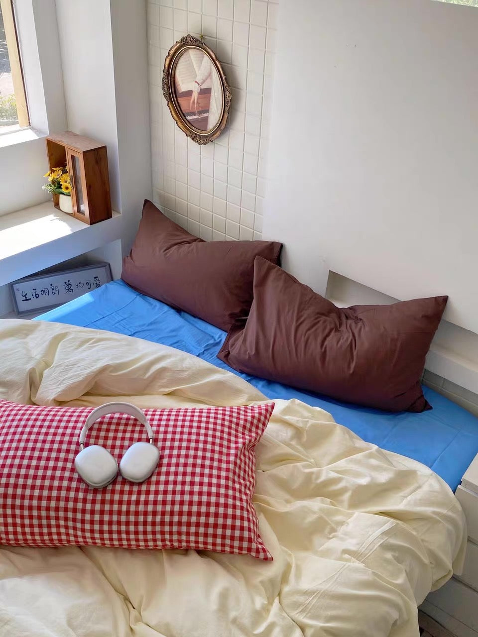 A cozy bedroom corner with a bed dressed in cream bedding, a bright blue sheet, and two brown pillows. A red-and-white gingham pillow with white headphones resting on it sits in the foreground.