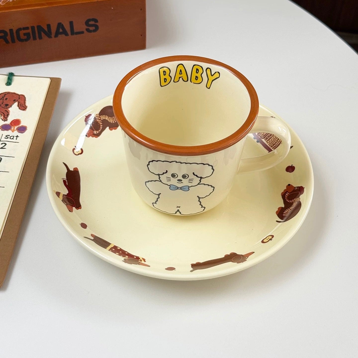 A cream-colored ceramic cup and saucer set on a white tabletop. The cup has a brown rim, yellow 'BABY' text, and a cartoon dog illustration.