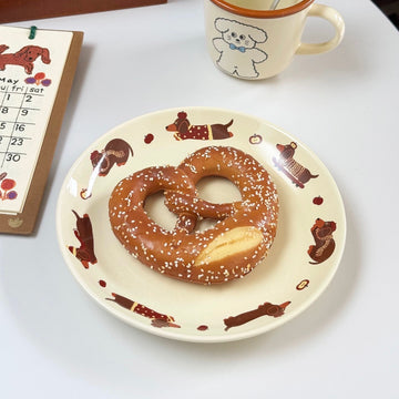 A pretzel cookie on a cream-colored ceramic plate decorated with brown dachshunds, set on a white table next to a mug and a calendar.