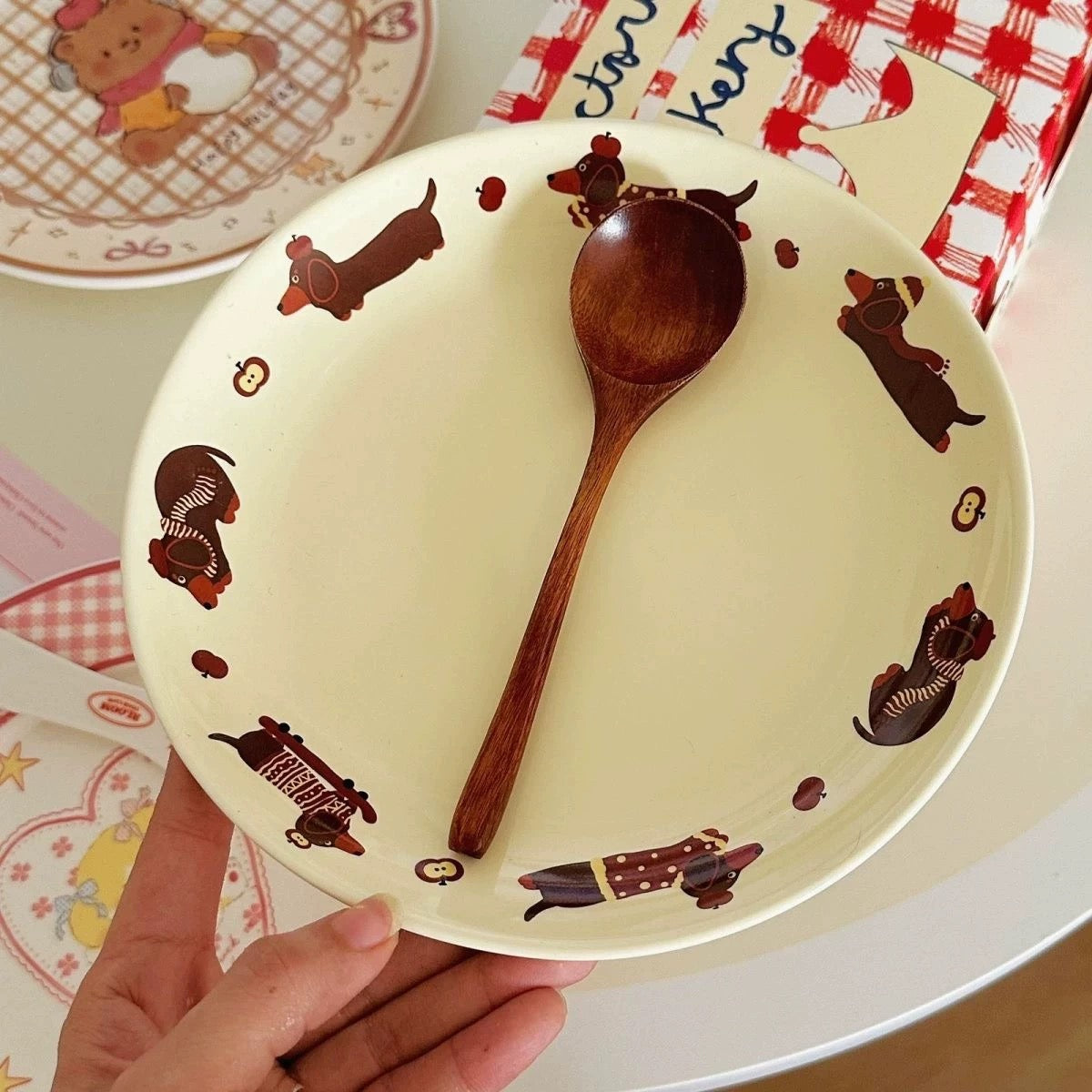 A hand holds a round cream-colored ceramic plate decorated with cartoon dachshund illustrations around the rim. A wooden spoon rests on the plate, and other styled table items are visible in the background.