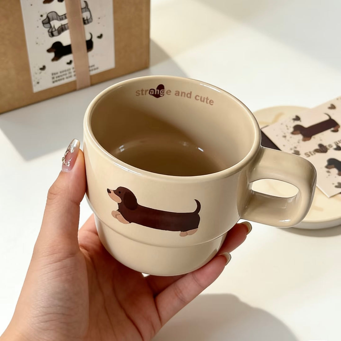 A hand with glittery nail polish holds a beige ceramic mug featuring a brown dachshund illustration and the text 'strange and cute' inside the mug.