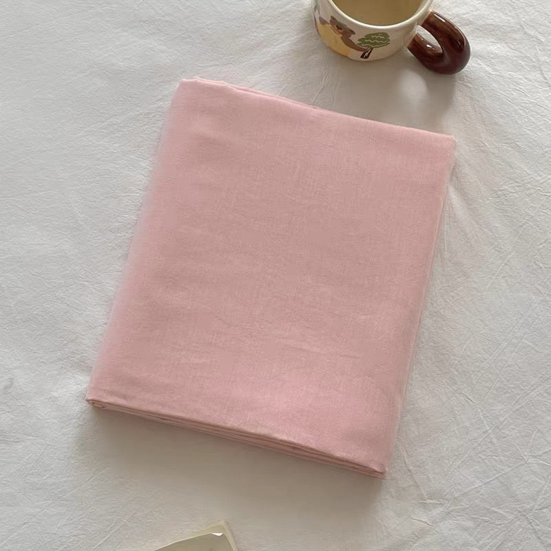 A folded light pink fabric item, likely a blanket or throw, displayed on a textured white fabric surface with a ceramic mug partially visible in the top right corner.