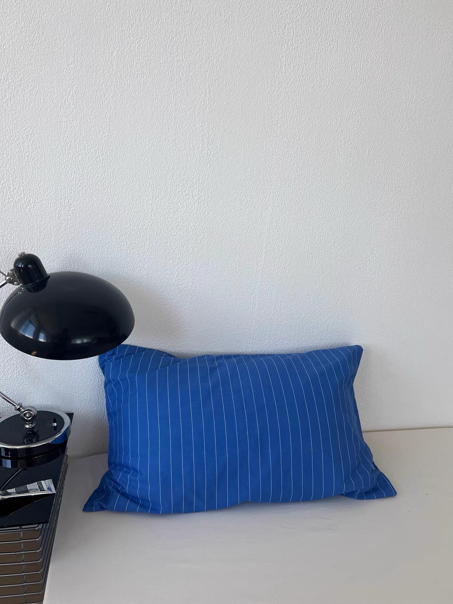 A rectangular blue throw pillow with thin white vertical pinstripes, resting on a white surface against a white wall next to a black desk lamp.