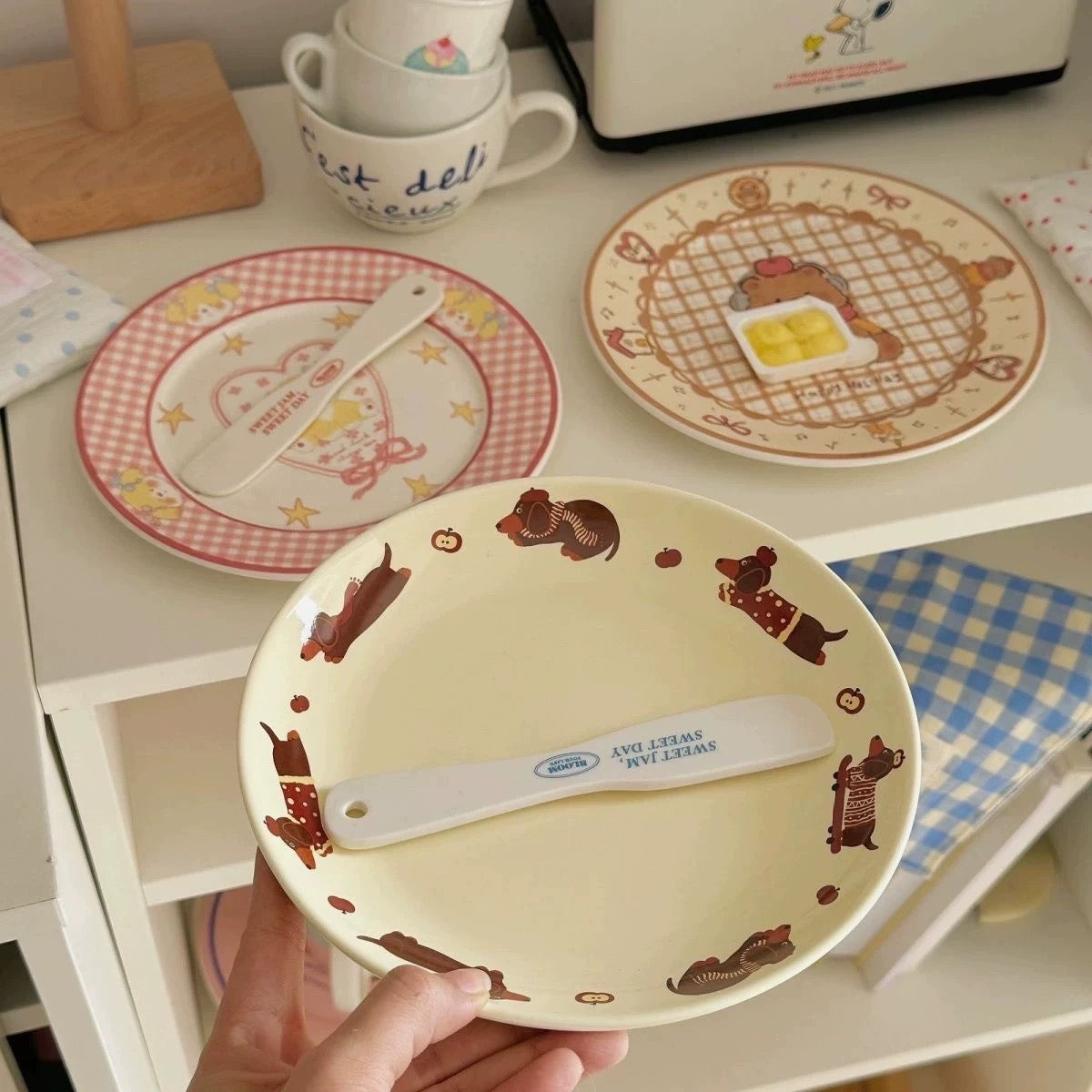 A hand holds a round cream-colored plate decorated with brown cartoon animals and a white spatula. In the background, other matching plates and cups sit on a white shelf.