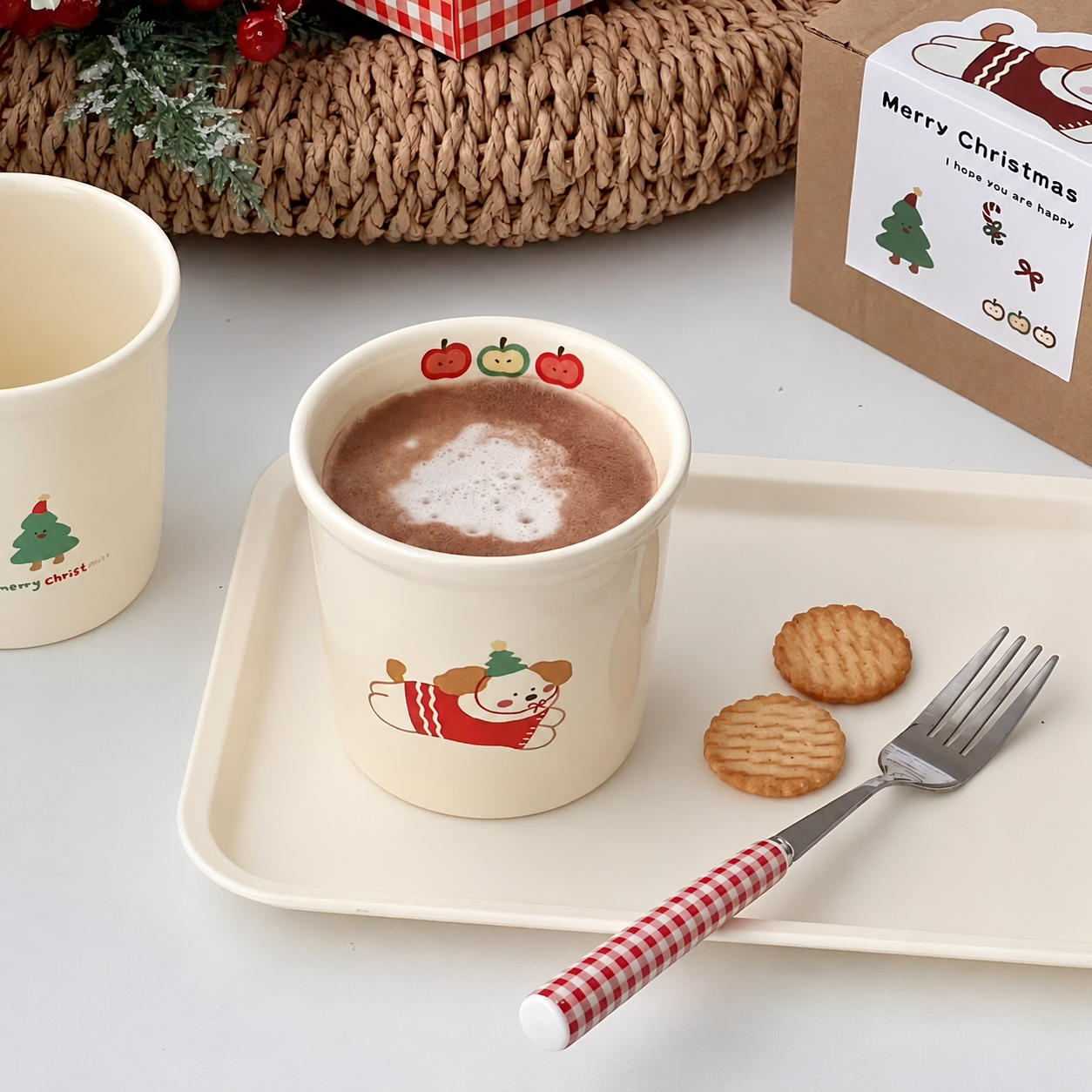 A festive cream-colored ceramic mug with a cartoon dog in a red sweater and pumpkin illustrations, filled with a hot chocolate drink on a white tray. A matching cookie cup, cookies, a fork, and a 'Merry Christmas' gift box are arranged around it on a white surface.