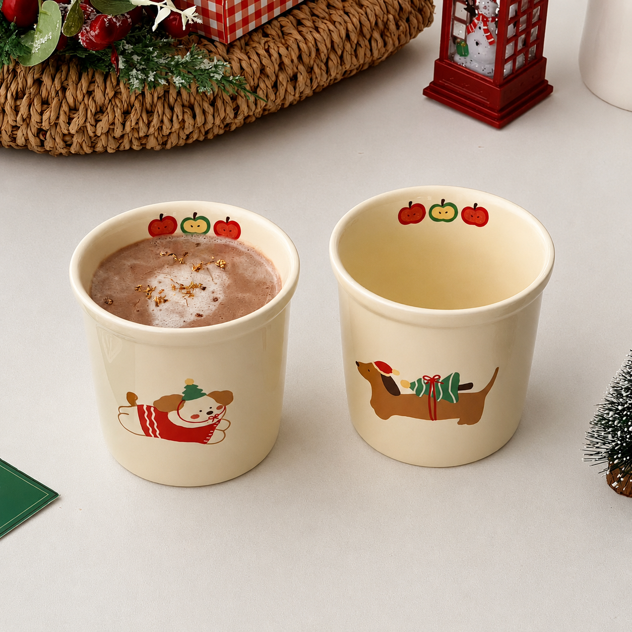Two cream-colored ceramic mugs with holiday illustrations, one filled with hot chocolate and the other empty, set on a light tabletop with Christmas decorations in the background.