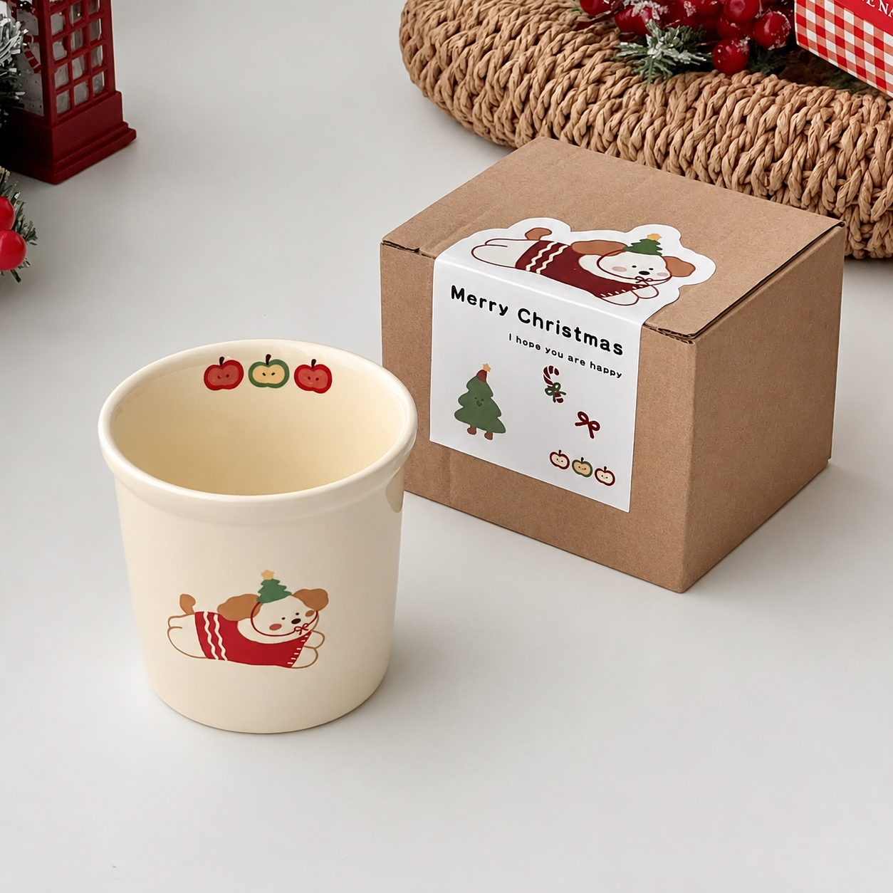 A cream-colored ceramic mug with a cute cartoon dog wearing a red sweater and green hat, shown next to its brown cardboard gift box decorated with a 'Merry Christmas' label.