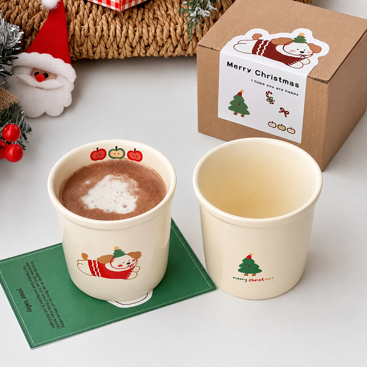 A Christmas-themed ceramic mug set shown with its packaging box. The set includes a cream-colored mug filled with hot cocoa on a green coaster, showing a cartoon bear wearing a red sweater and green hat, and a second empty mug next to it.