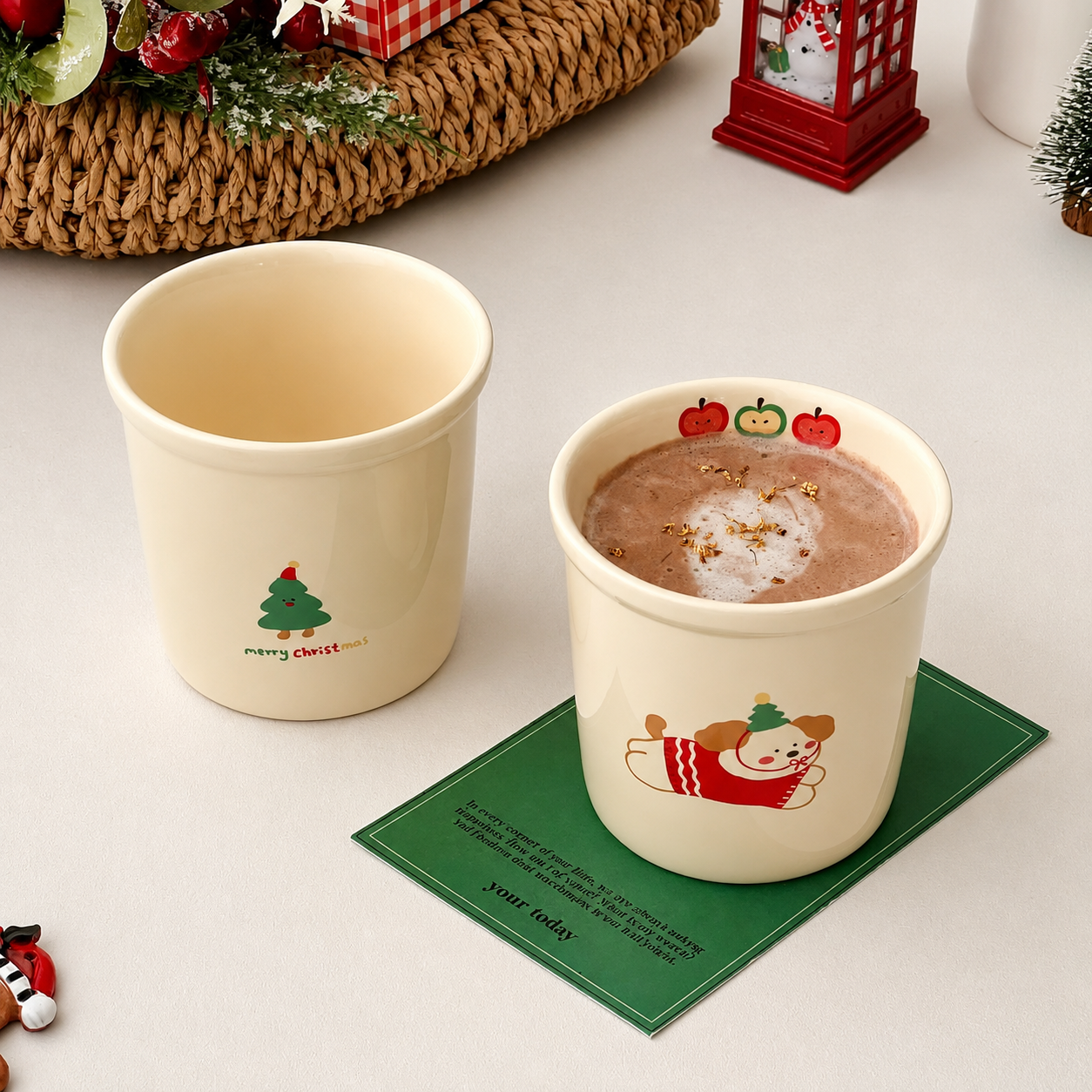 Two cream-colored ceramic cups with Christmas-themed illustrations, one empty and one filled with a hot chocolate drink and placed on a green coaster, set on a light tabletop with holiday decorations in the background.