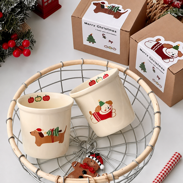 Two cream-colored ceramic Christmas mugs with dachshund and bear illustrations, displayed in a wire basket with kraft gift boxes in the background.