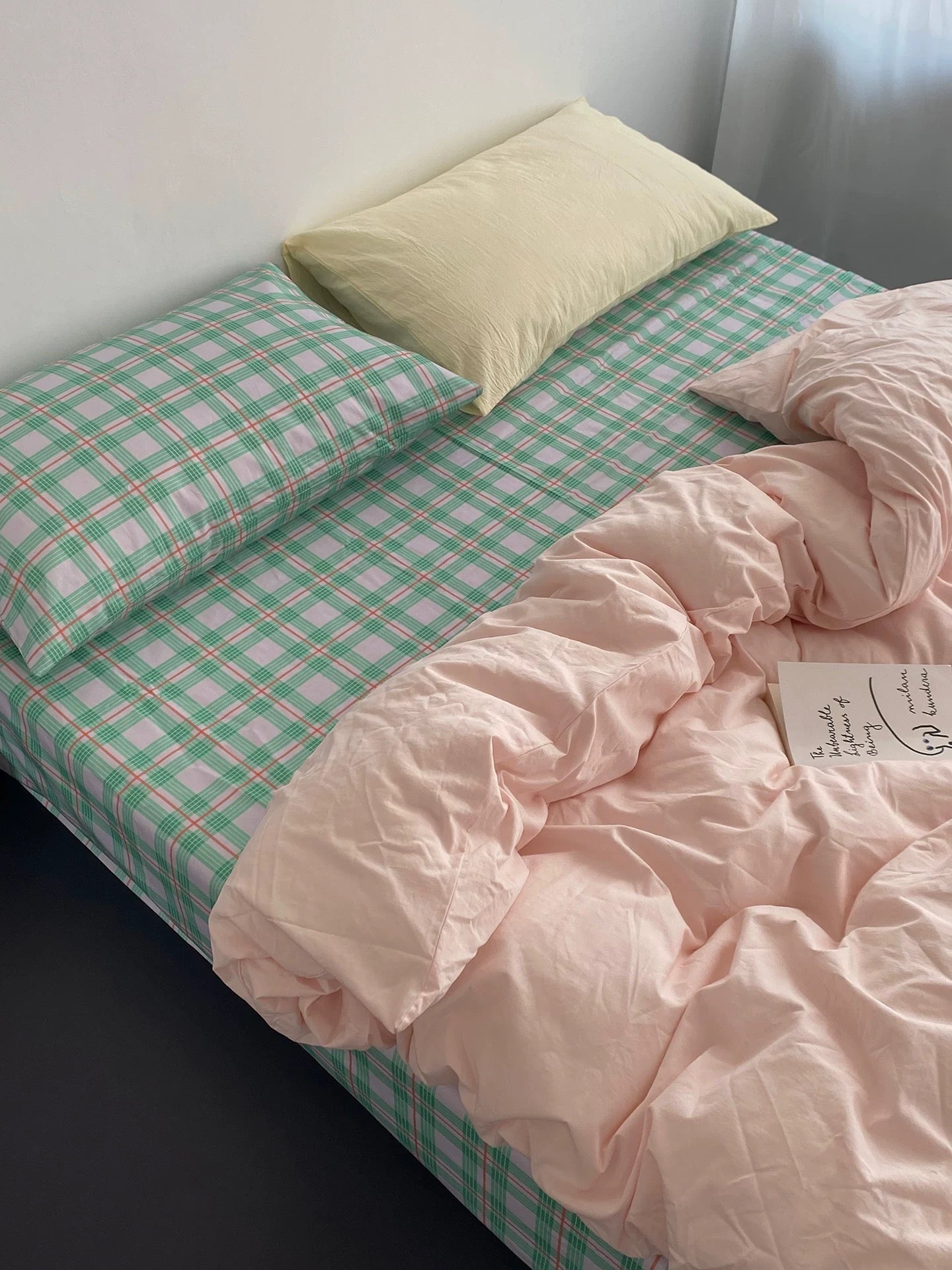 Bed with green checkered sheets and pink comforter against a white wall.