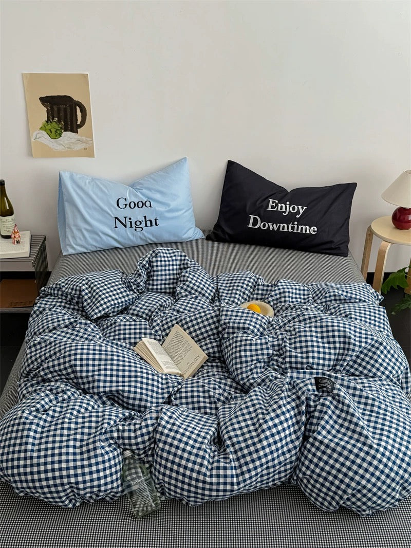Bed with checkered duvet, pillows labeled 'Good Night' and 'Enjoy Downtime', and a small table with a bottle and vase.