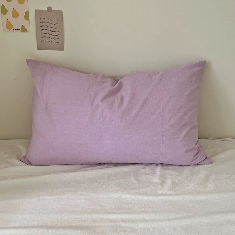 Plain muted lavender cotton pillowcase resting against cream walls on a white bed.