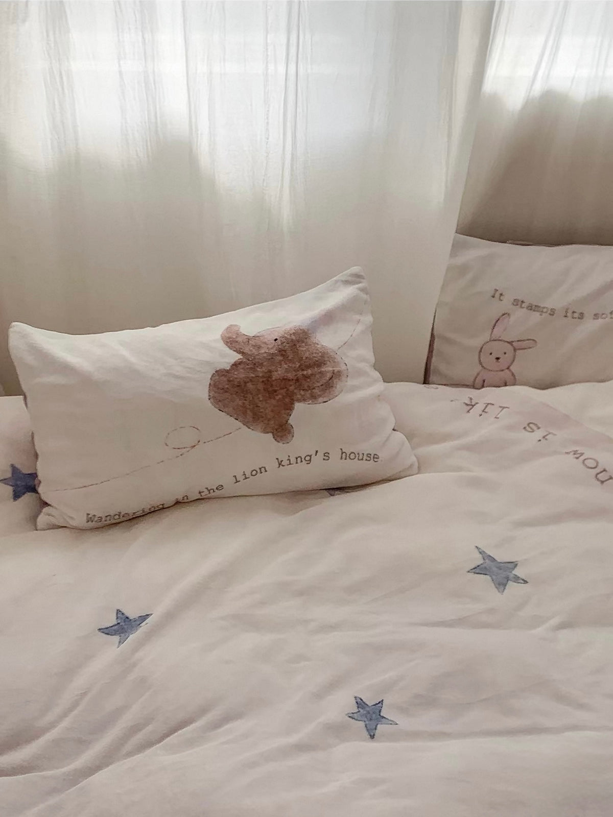 An elephant pillowcase rests above the pale star-covered bed near a sheer curtain.