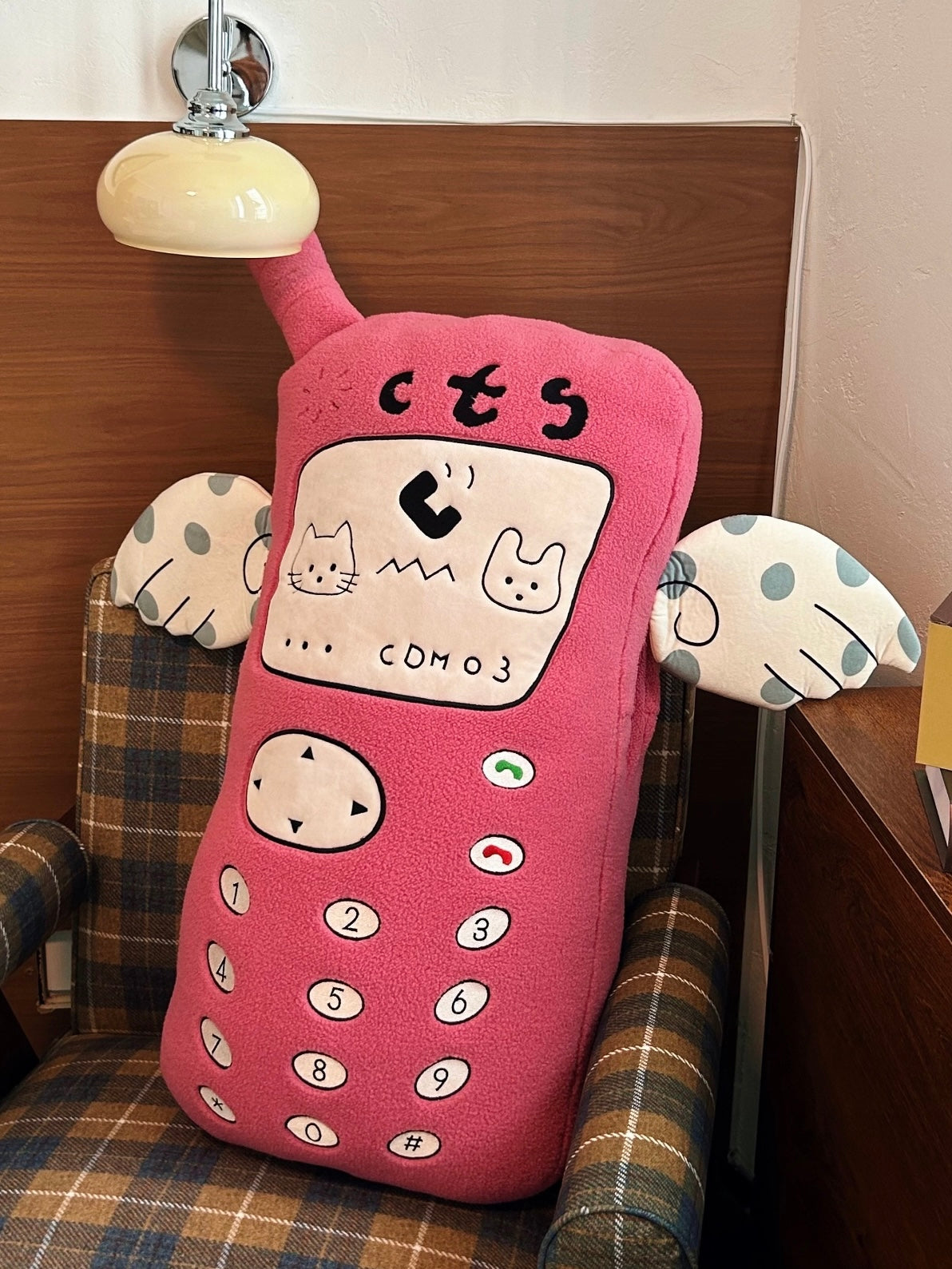 An embroidered phone pillow stands against a checked chair, with its pink antenna above the cartoon screen.