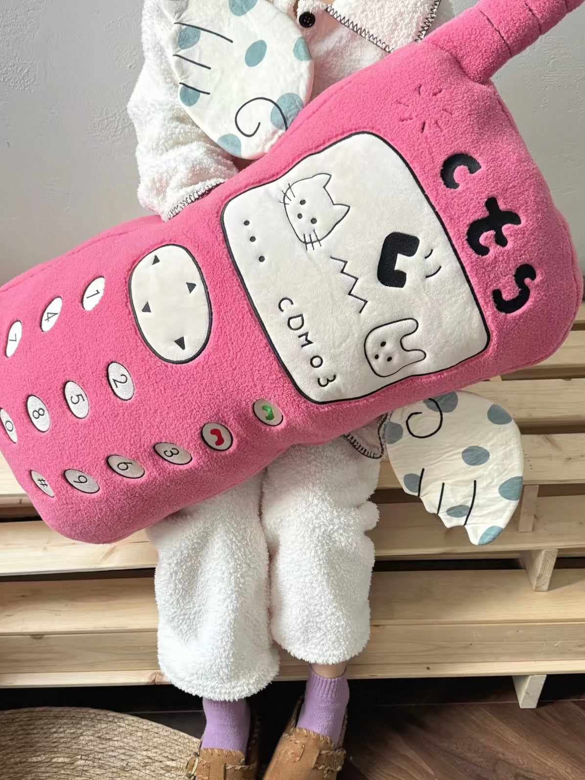 A pink embroidered telephone pillow is held at an angle, showing its cartoon screen, number buttons and antenna.