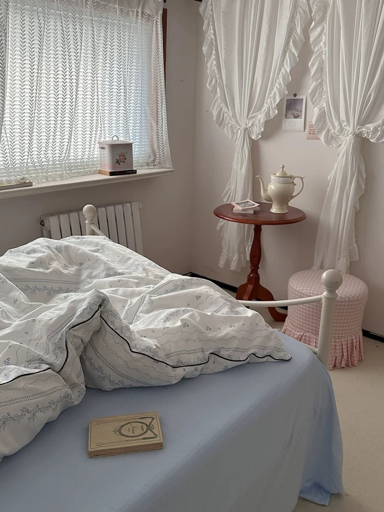 Bedroom with white bedding, a small table with a teapot, and a window with white curtains.