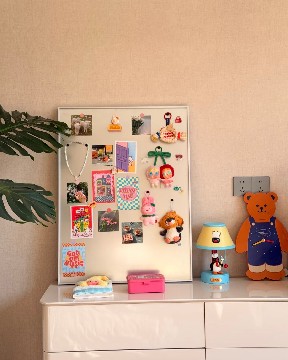 A white rectangular magnetic board mounted on a beige wall above a white cabinet, decorated with photos, cards, a stethoscope, and small hanging accessories. A potted plant is on the left, and a small lamp and a teddy bear figure are on the right.