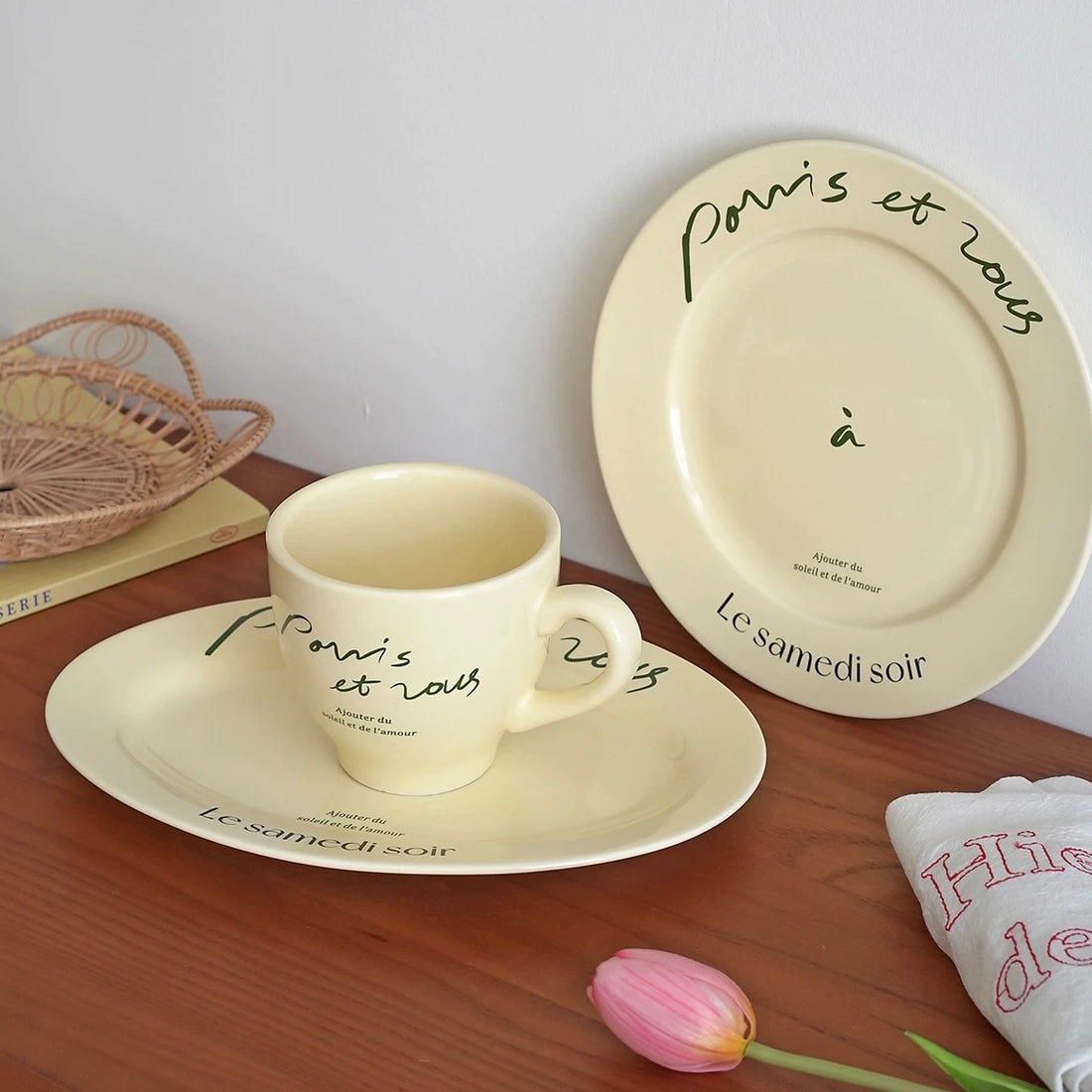 Cream porcelain mug and round plate with black French script displayed together on wooden tabletop
