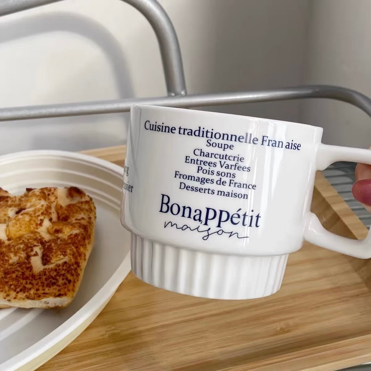 A white ceramic French coffee mug with 'BonaAppétit maison' printed in navy blue text, held by a hand over a wooden tray with toast in the background.