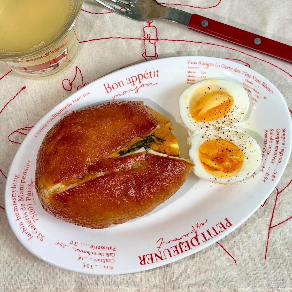A breakfast plate from Petit Défonder featuring a golden-brown bagel sandwich with cheese and greens, served with two halves of a hard-boiled egg with runny yolks. The white plate has red French text and illustrations, set on a patterned beige tablecloth next to a glass of juice and a knife.