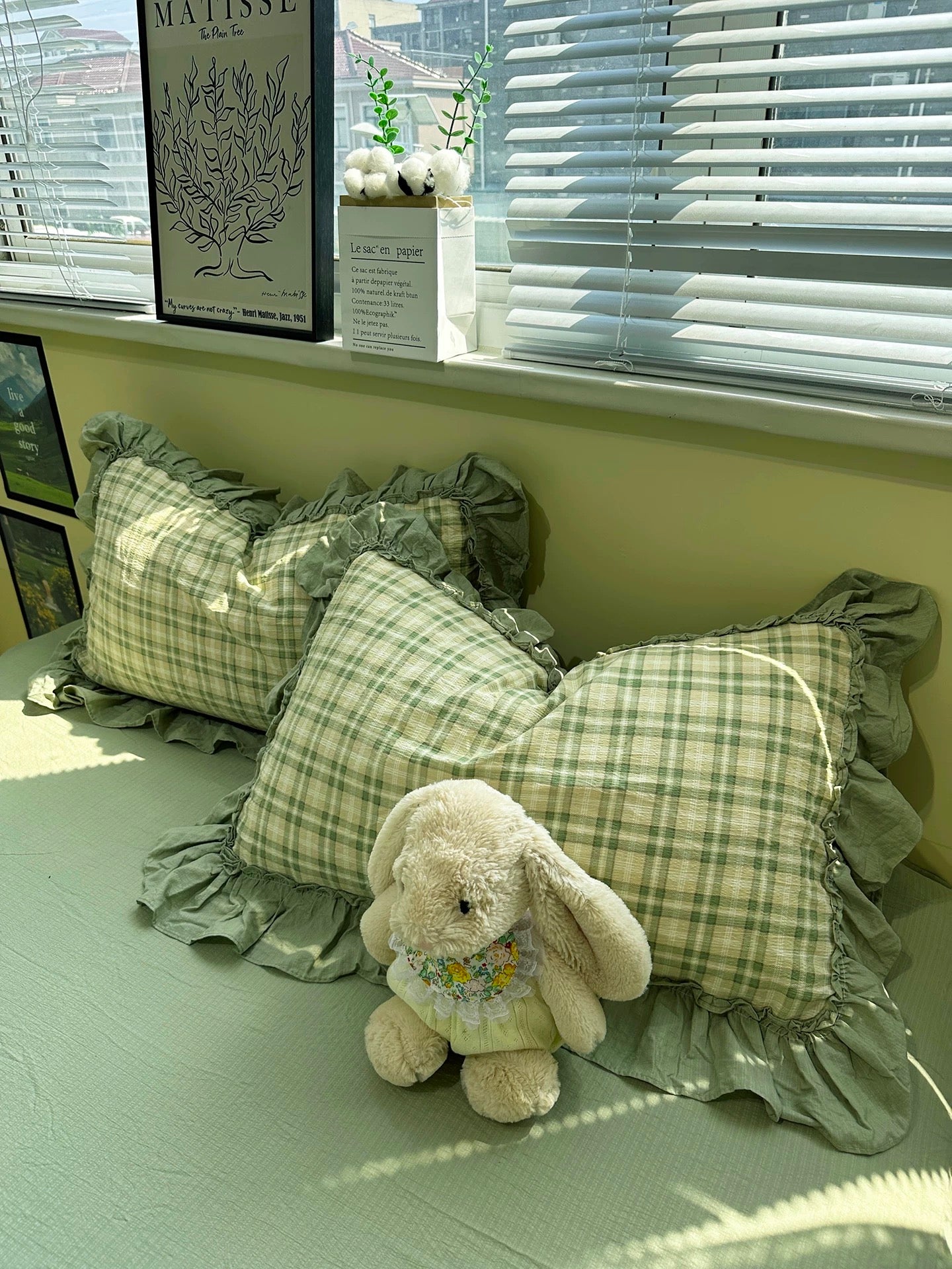 Plaid pillows with ruffles and a plush toy on a bed in a room with a window and decor.