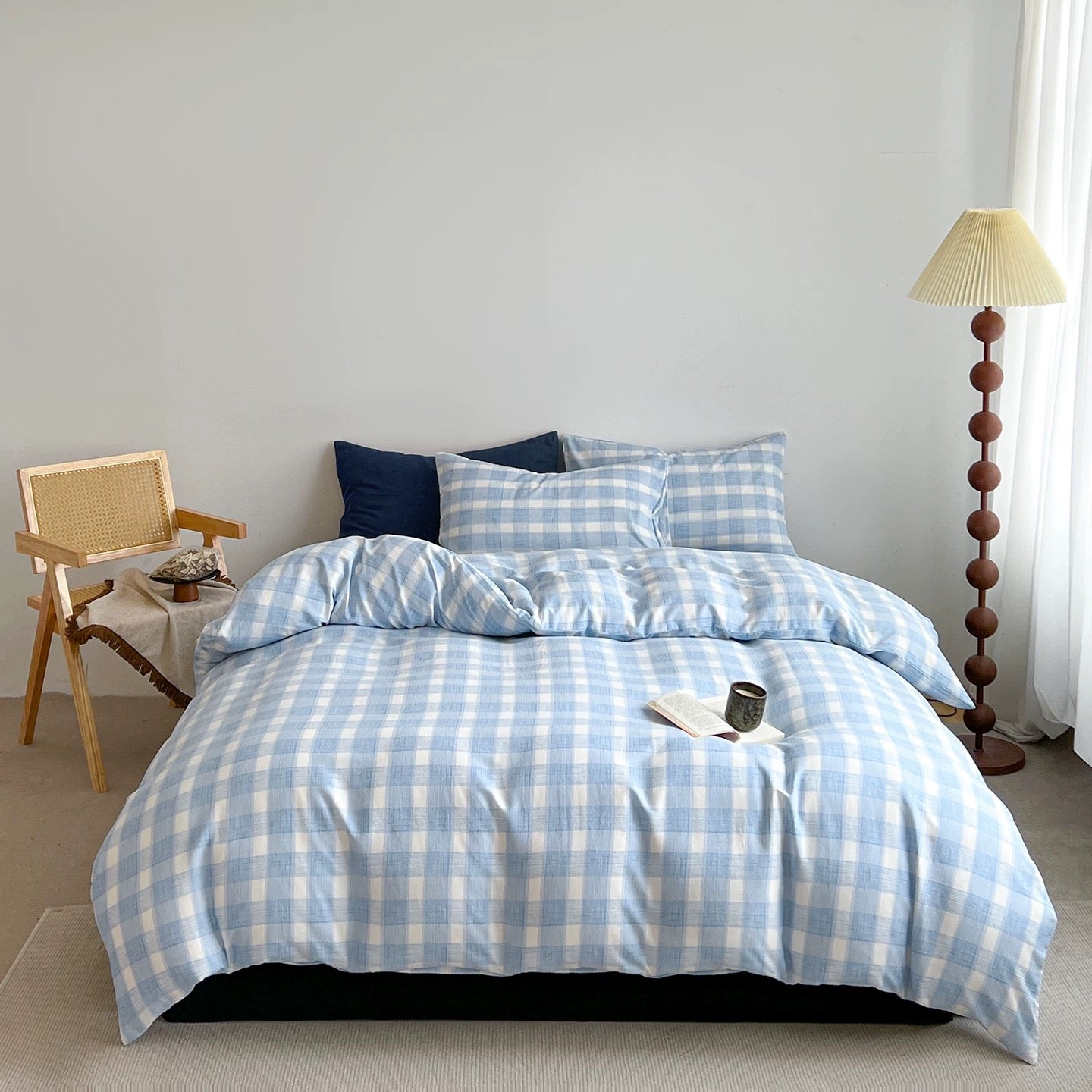 A bed dressed in a light blue and white gingham checkered bedding set, styled in a bright bedroom with a wooden chair, floor lamp, and white curtains.