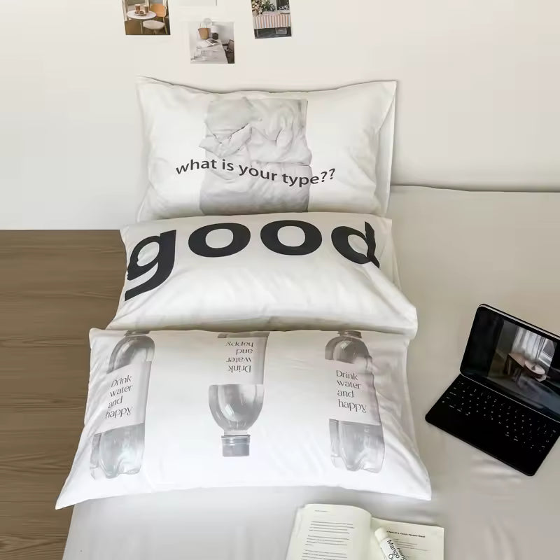 A styled bed setup featuring three white printed pillows with black text and graphic designs, alongside a black laptop on a light wood floor against a white wall.