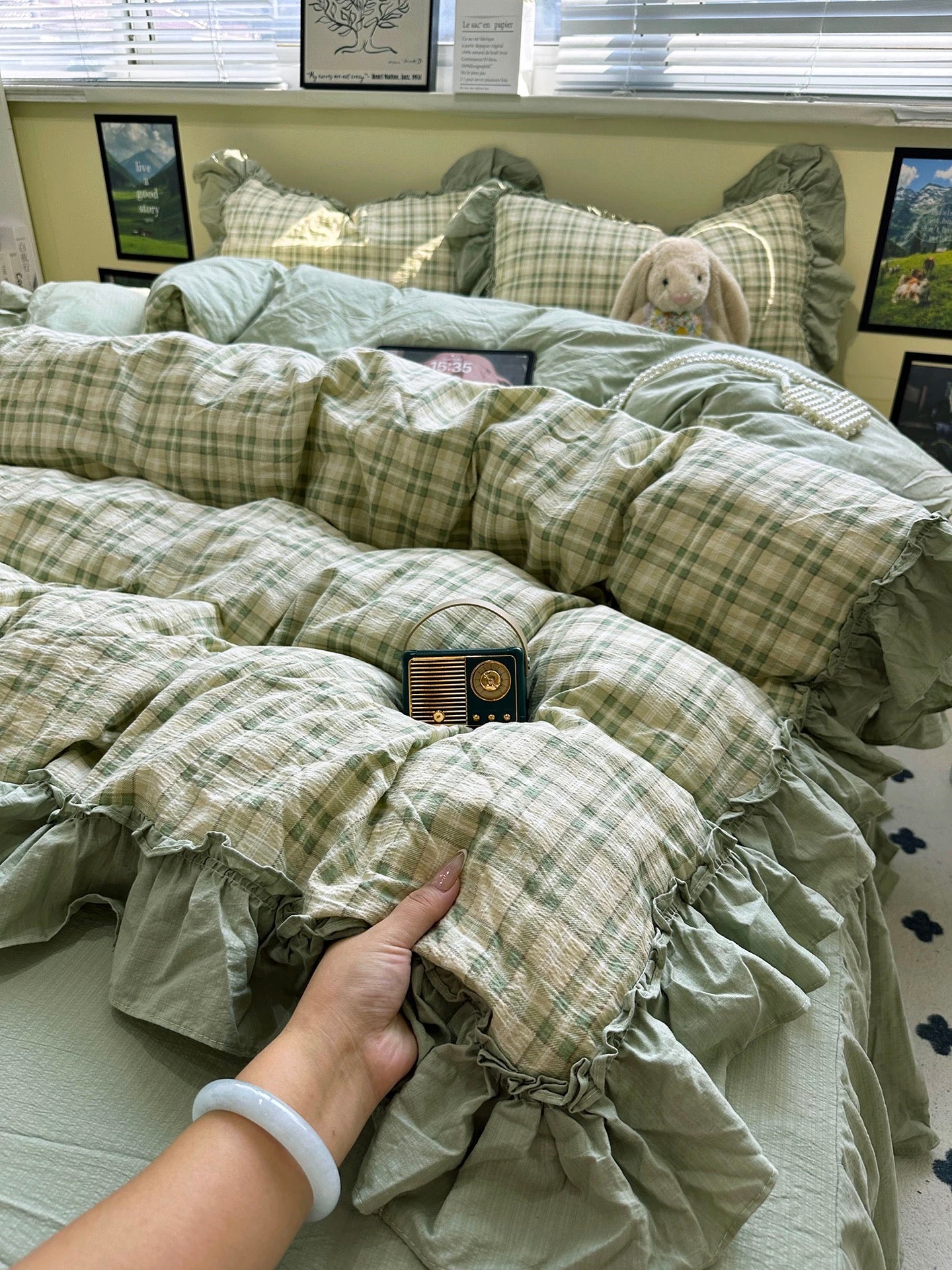 Green plaid comforter on a bed with a hand touching it, in a room with framed pictures on the wall.