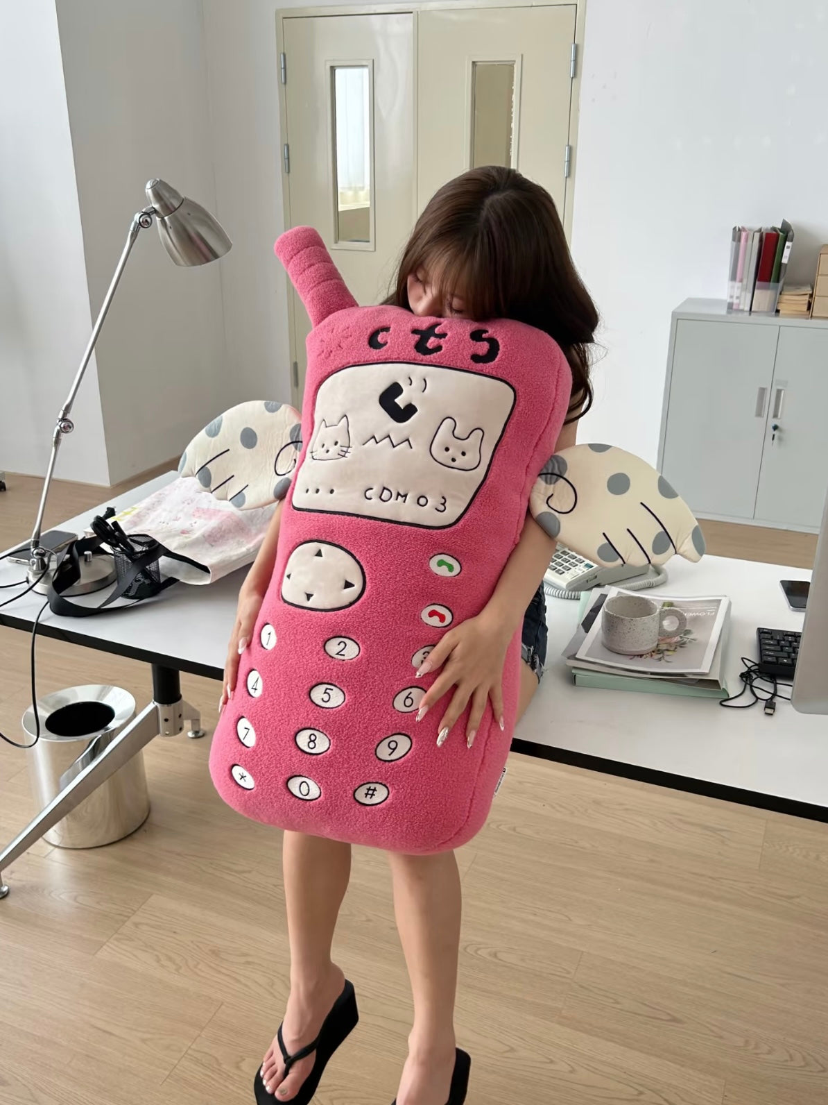 A pink phone hand warmer pillow is held upright, showing an embroidered keypad and spotted fabric arms.