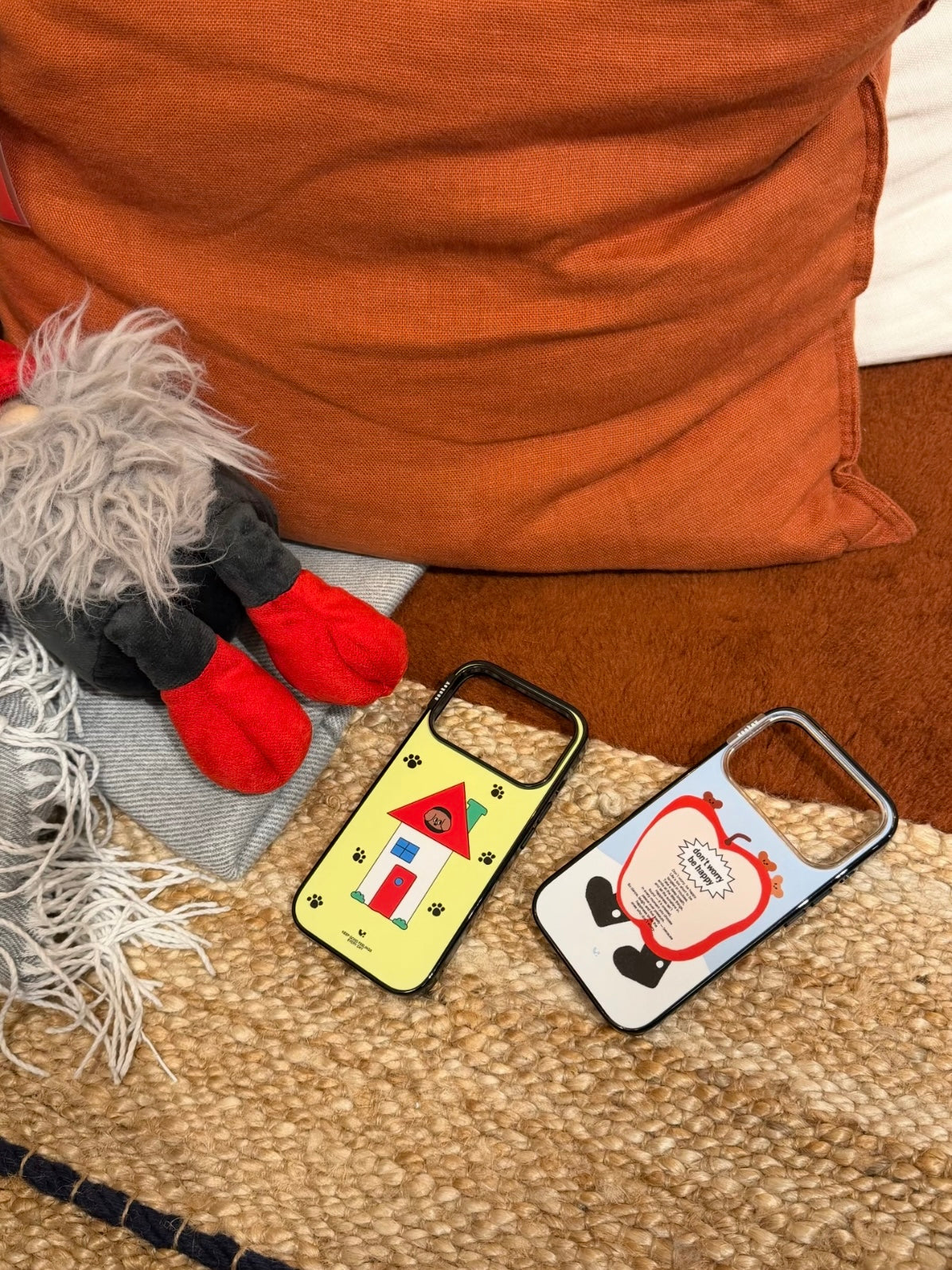 Two illustrated phone case designs rest on a chair, with a red apple on blue and a puppy house on yellow.