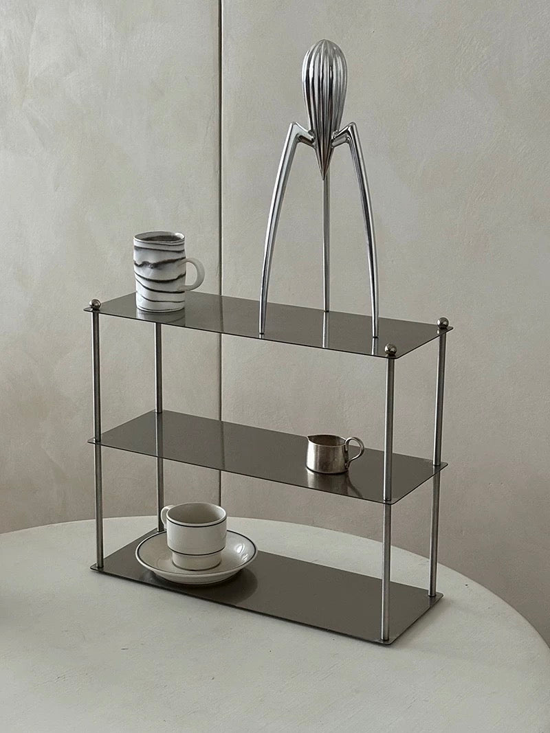 Metallic juicer on a glass shelf with cups and saucers below against a neutral background