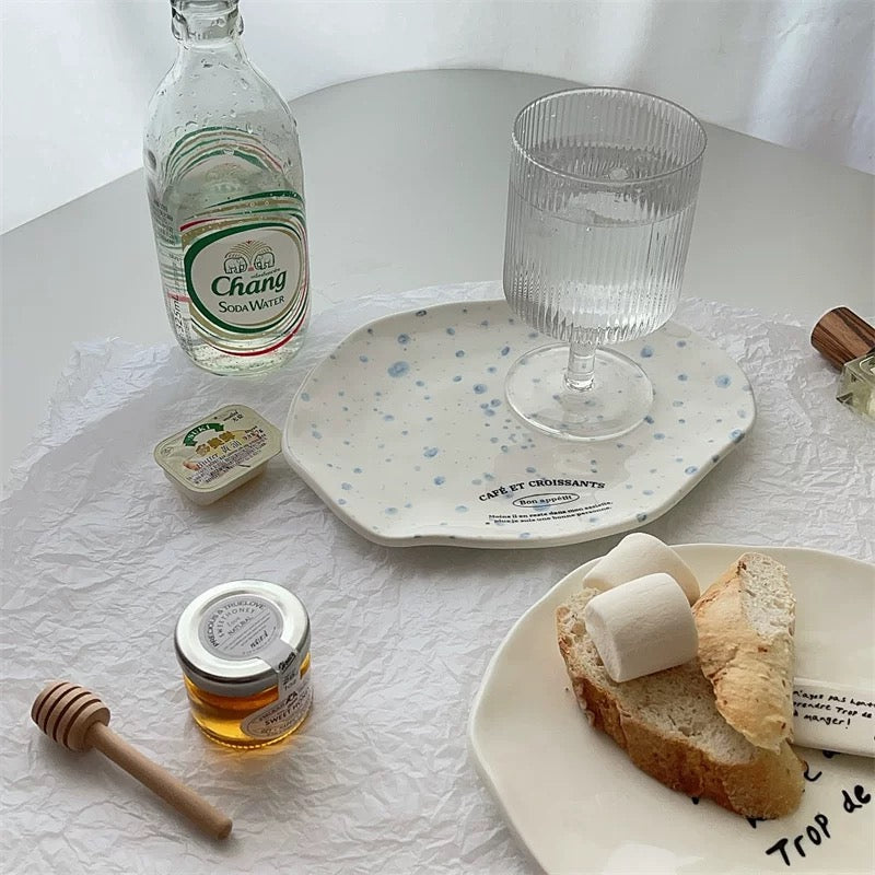 Two ceramic plates with spoons, styled on a crumpled white paper background with bread, a bottle of perfume, and small jars as props.