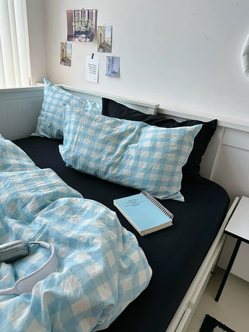 Bed with blue checkered bedding and pillows, book, and remote control on a white wall background.