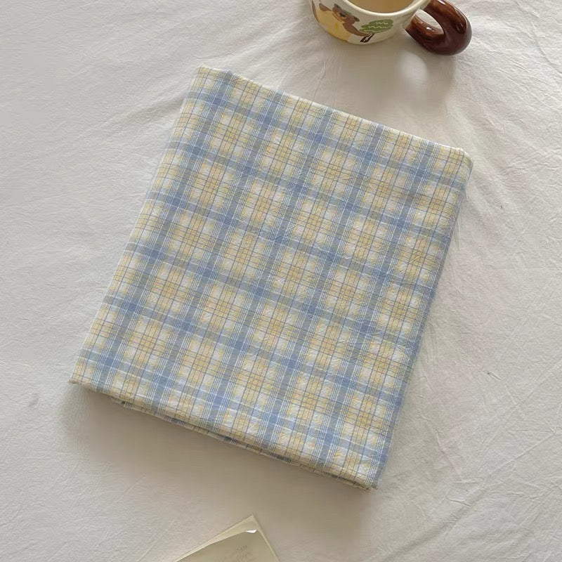A folded plaid fabric item, likely a blanket or throw, in a soft blue, yellow, and white checkered pattern, styled on a white textured blanket with a ceramic mug partially visible at the top.