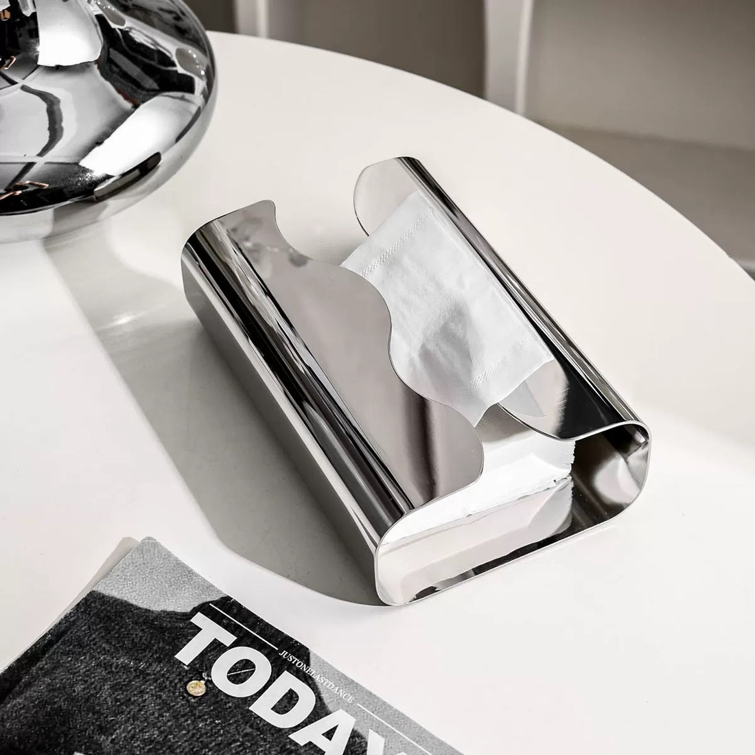 Silver tissue holder with tissues on top of a magazine in a minimalistic setting.