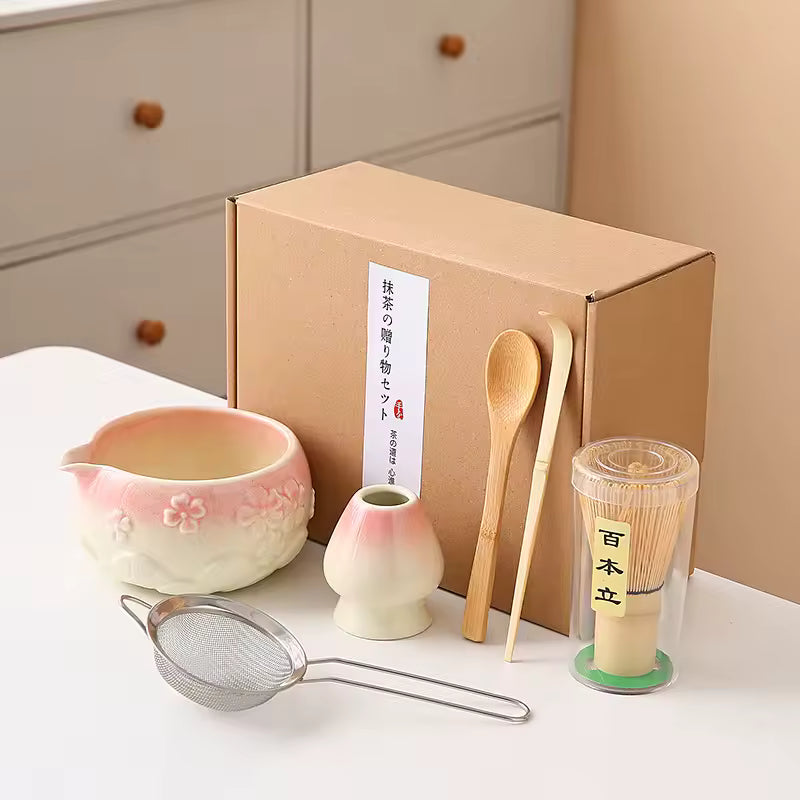 Tea-making set with pink bowl, small vase, wooden spoon, and other tools in front of a cardboard box.