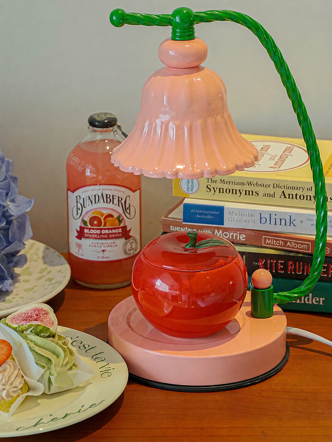 A retro-style pink and green table lamp shaped like a bell-shaped shade, a red apple, and a green stand, displayed on a wooden table next to a bottle of Sangdaberg blood orange soda and a stack of books.
