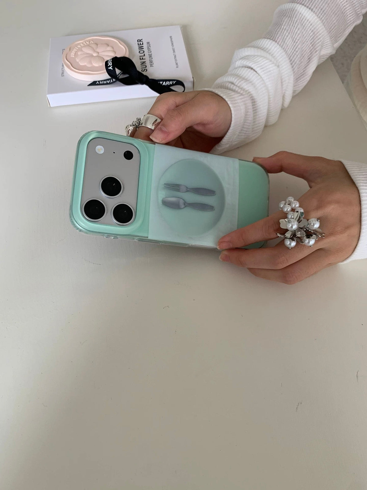 A mint green phone case held sideways in one hand, showing the plate and cutlery artwork on the back.