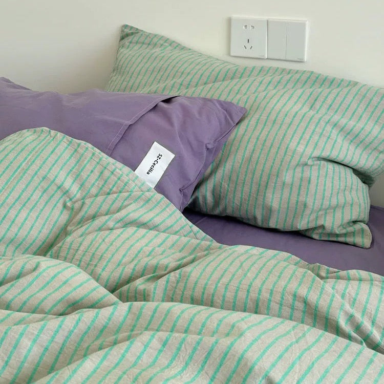 Bed with green and white striped bedding and purple pillows against a white wall.